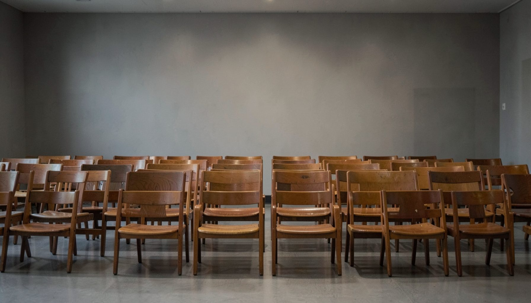 A row of empty wooden chairs facing forward in a plain meeting hall, muted gray and warm brown tones, quiet and reflective
